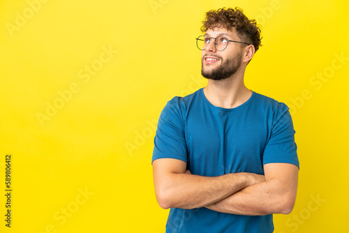 Young handsome caucasian man isolated on yellow background looking up while smiling