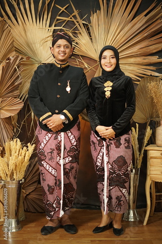 A couple in traditional dress pose for a photo in front of a large wall with a palm tree behind them.