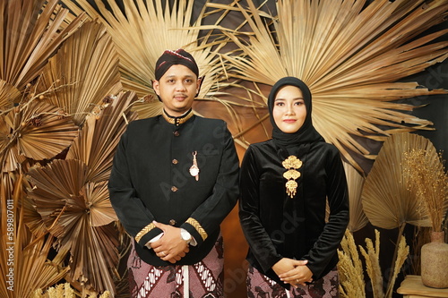 A couple in traditional dress pose for a photo in front of a large wall with a palm tree behind them.