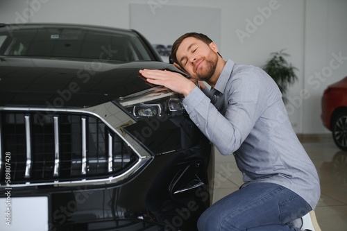 Visiting car dealership. Handsome bearded man is stroking his new car and smiling.