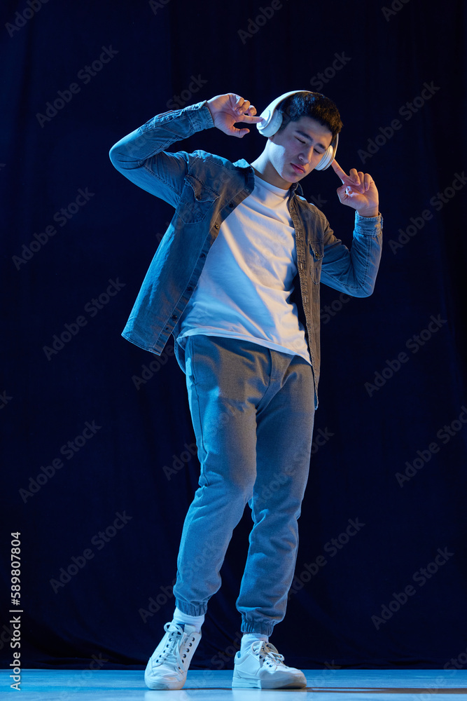Full-length image of young guy in casual clothes listening to music in headphones against dark background in neon light. Enjoying sounds. Concept of human emotions, youth, fashion, lifestyle Full-length image of young guy in casual clothes listening to music in headphones against dark background in neon light. Enjoying sounds. Concept of human emotions, youth, fashion, lifestyle