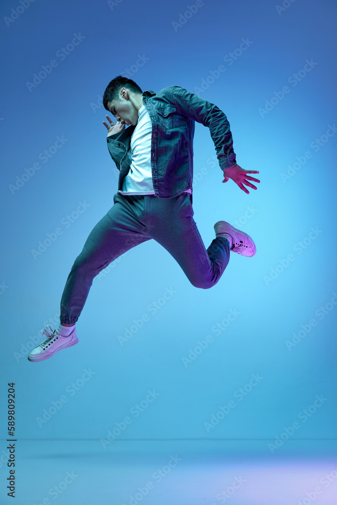 Delightful, reaching success. Full-length image of excited young man in casual clothes jumping against blue background in neon light. Concept of human emotions, youth, fashion, lifestyle
