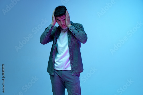 Making mistakes. Portrait of young guy holding head with hands with sad, stressful face against blue background in neon light. Concept of human emotions, youth, casual fashion, lifestyle