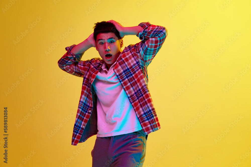 Astonished look. Shocked and surprised. Young guy in casual stylish clothes showing emotions against yellow background in neon light. Concept of human emotions, youth, fashion, lifestyle Astonished look. Shocked and surprised. Young guy in casual stylish clothes showing emotions against yellow background in neon light. Concept of human emotions, youth, fashion, lifestyle
