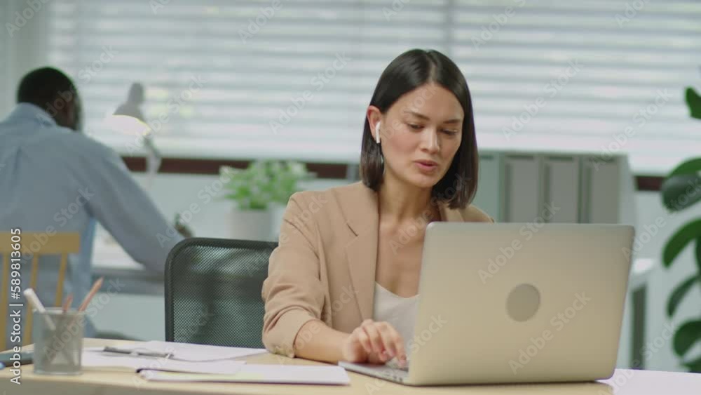 Medium shot of beautiful businesswoman in formalwear and wireless earphones having discussion via web call on laptop while working in office Medium shot of beautiful businesswoman in formalwear and wireless earphones having discussion via web call on laptop while working in office