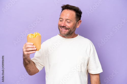 Middle age caucasian man holding fried chips isolated on purple bakcground looking to the side and smiling © luismolinero