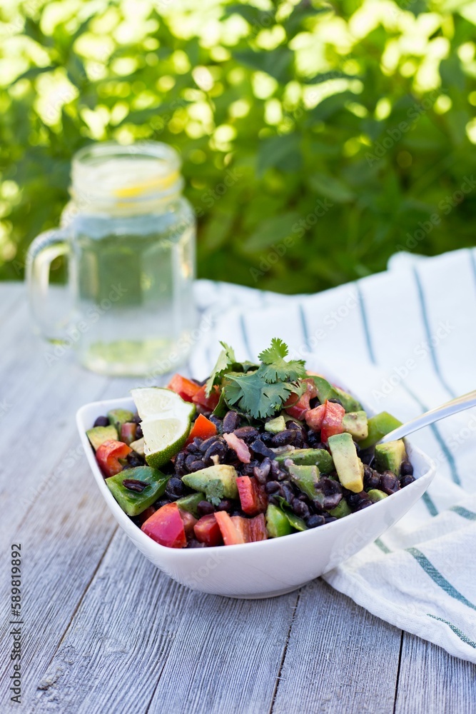 Bowl of healthy salad with lime, avocados, tomatoes, and beans with lemonade water on the gray table