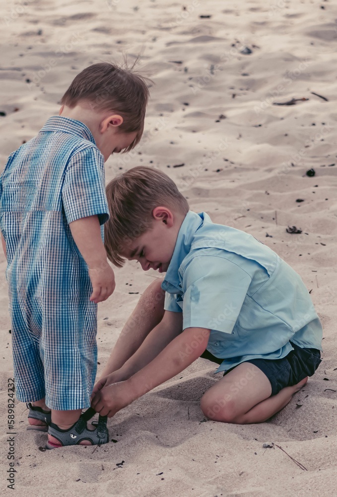 Boy helping his brother to put on his shoes on a sandy beach Boy helping his brother to put on his shoes on a sandy beach