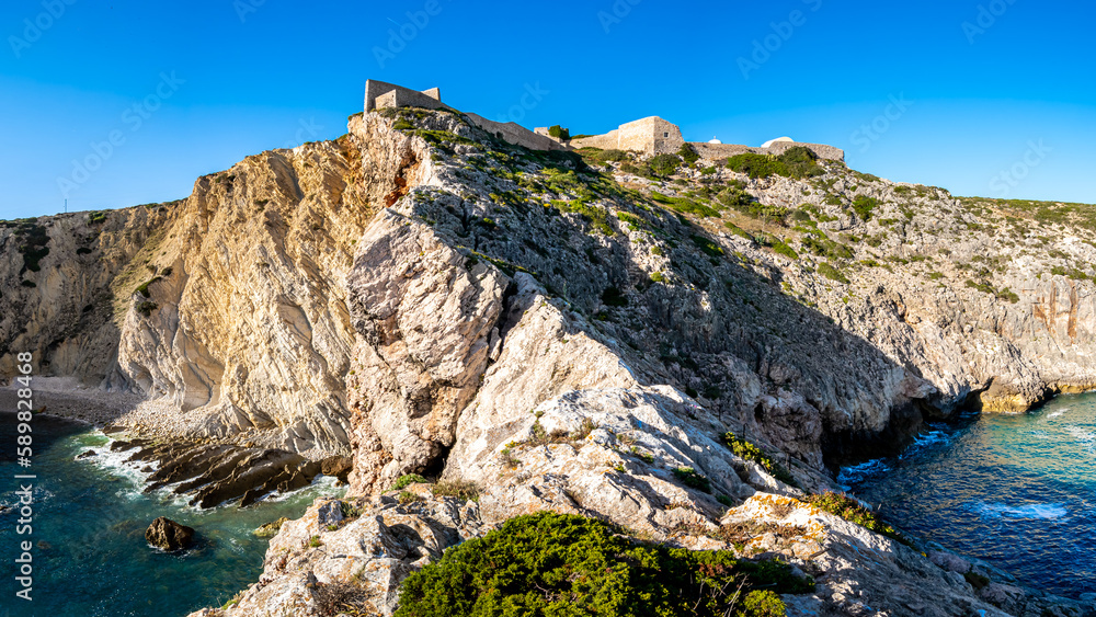 A breathtaking panoramic view captures the rugged beauty of a ragged cliff with the ruins of fort Fortaleza de Belixe atop, seen from a narrow ridge with abyss left and right nearby Sagres, Portugal.