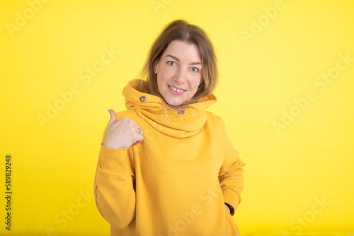 Charming 40-year-old woman in yellow hoodie on yellow background looks at camera, pointing with her hand OK. Sports.  © Татьяна Волкова