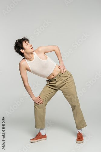 full length of trendy pansexual person in crop top and beige pants looking away on grey background.