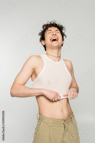 excited bigender person pulling down crop top and laughing with closed eyes isolated on grey.