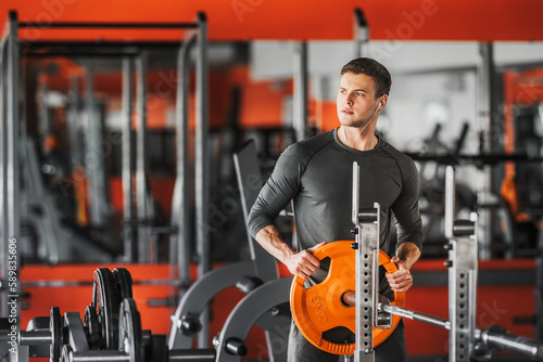 Young cross fit athlete preparing barbell for lifting weight at the gym. Practicing powerlifting. A man holds a disc from a barbell weighing 20 kg. Copy space.