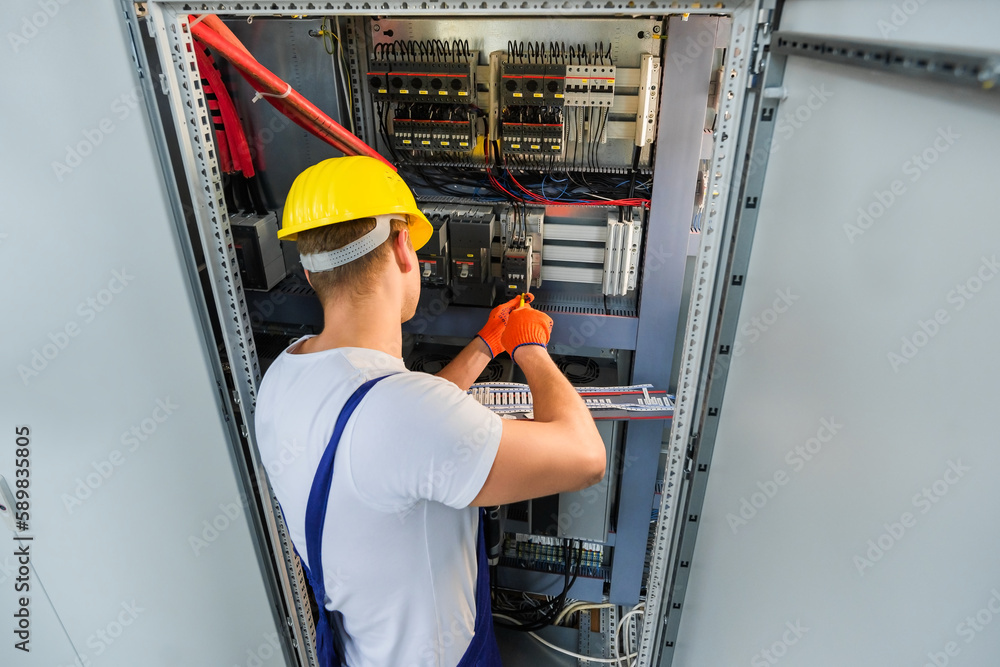 Electrician with screwdriver tighten up switching electric actuator equipment in fuse box. View from the back, a young specialist in a yellow hard hat and overalls. Indoor. Electrician with screwdriver tighten up switching electric actuator equipment in fuse box. View from the back, a young specialist in a yellow hard hat and overalls. Indoor.