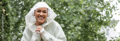 smiling african american woman in waterproof raincoat and wireless headphones standing under rain, banner.
