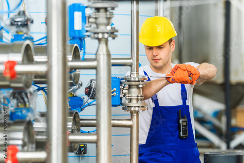 Fotografie A young factory worker in yellow hard hat tightens nut with wrench on connection of stainless steel pipes