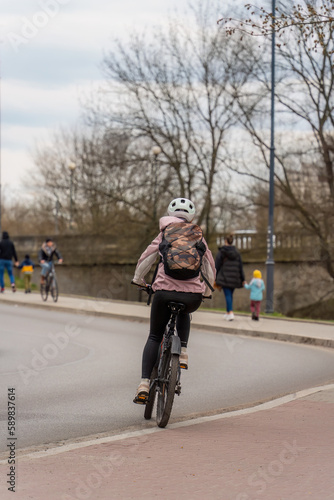 Fototapeta Naklejka Na Ścianę i Meble -  A girl rides a bike in a jacket with a backpack behind her back in a white helmet along the city street on a cloudy day in early spring, eco sustainable urban transport, street photography