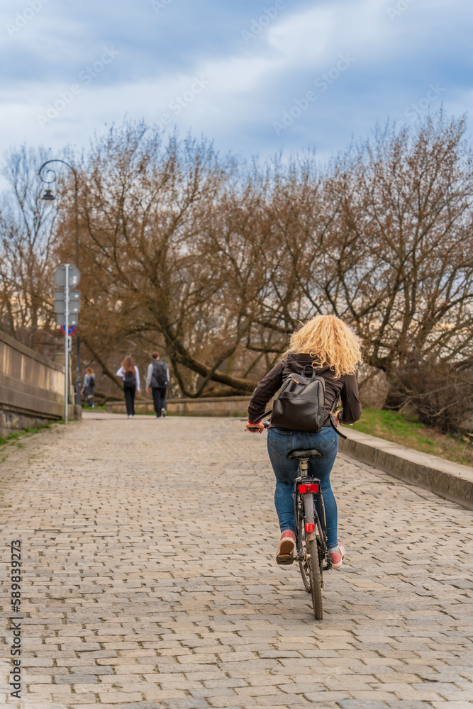 A blonde with long hair rides a bicycle in a jacket, jeans with a backpack on her back along the city street on a cloudy day in early spring, eco sustainable urban transport, street photography