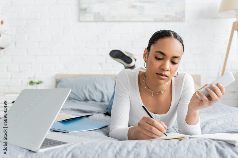 Obraz premium African american student holding smartphone and writing on notebook near laptop on bed.