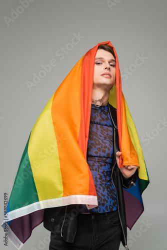 trendy pangender person covering himself with rainbow flag and looking at camera isolated on grey. photo