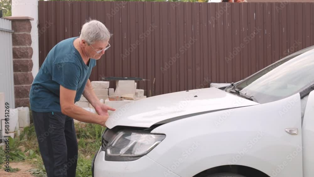 The gray-haired man cannot open the hood of the car. The hood is jammed and the man cannot open it. The gray-haired man cannot open the hood of the car. The hood is jammed and the man cannot open it.