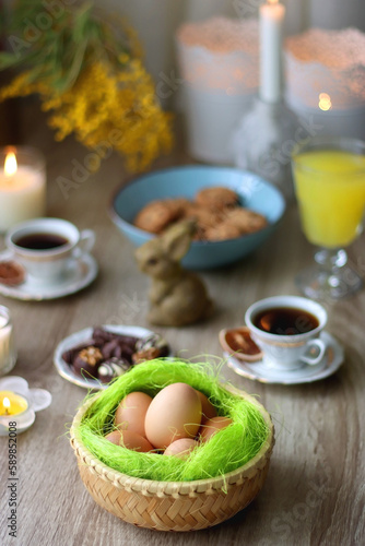 Easter eggs in the basket, bowl of cookies, chocolate pralines, Easter bunny figurine, cups of tea, glasses of juice, flowers and lit candles on the table. Selective focus.