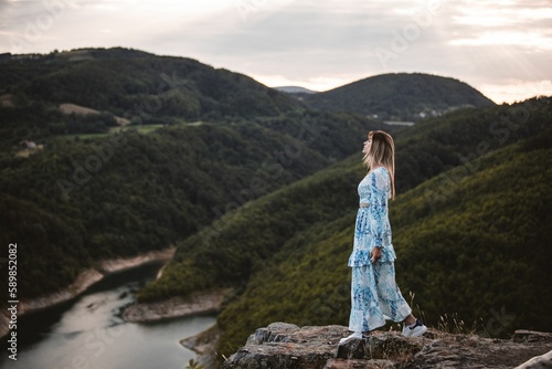 Beautiful Caucasian girl in a dress standing on a rock enjoying the sunset over the river and hills.