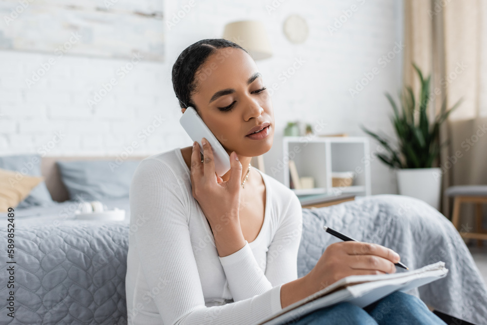 pretty african american student taking notes while talking on smartphone and studying form home. pretty african american student taking notes while talking on smartphone and studying form home.