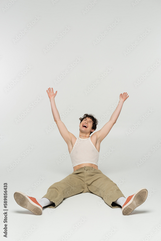 excited nonbinary person in beige pants laughing while sitting with raised hands on grey background. excited nonbinary person in beige pants laughing while sitting with raised hands on grey background.