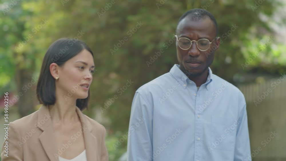 Medium shot of Caucasian businesswoman speaking with black male coworker while walking together along street on summer day Medium shot of Caucasian businesswoman speaking with black male coworker while walking together along street on summer day
