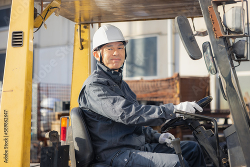 Middle-aged Asian male worker with forklift on factory grounds.
