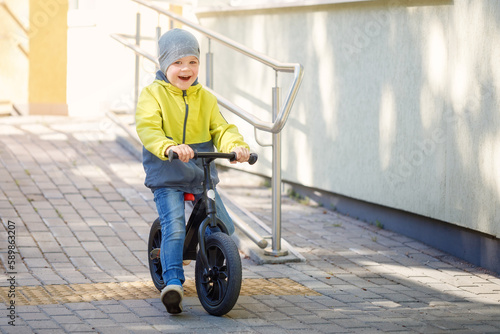 A happy little boy in a yellow jacket and a gray hat rides downhill on his new balance bike in spring time