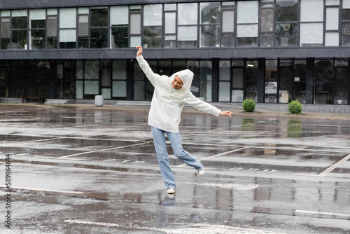 full length of happy african american woman in waterproof raincoat and jeans having fun during rain.