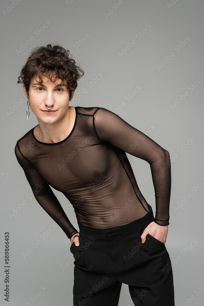 trendy bigender person in black transparent top posing with hands in pockets isolated on grey. trendy bigender person in black transparent top posing with hands in pockets isolated on grey.