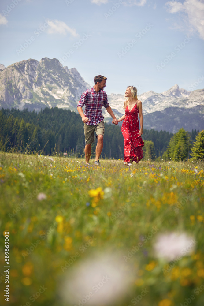 Beautiful young couple holding hands and running on green meadow. Mountains in background. Fun, togetherness, lifestyle, nature concept.