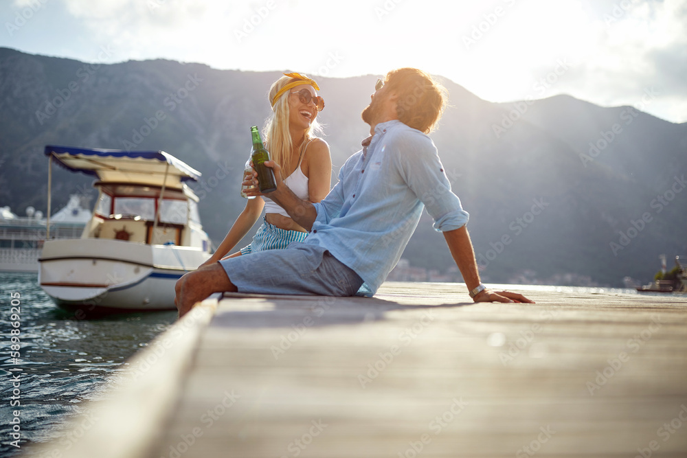 A young happy couple is sitting on the dock on the seaside and chatting with a drink. Love, relationship, holiday, sea