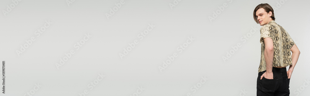 happy nonbinary person holding hands in back pockets and smiling at camera isolated on grey, banner. happy nonbinary person holding hands in back pockets and smiling at camera isolated on grey, banner.