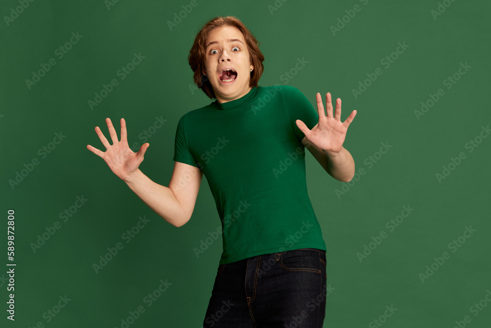 Portrait with young frightened man , guy screaming and raising hands with fear face over green studio background. Horror, thriller, scare story