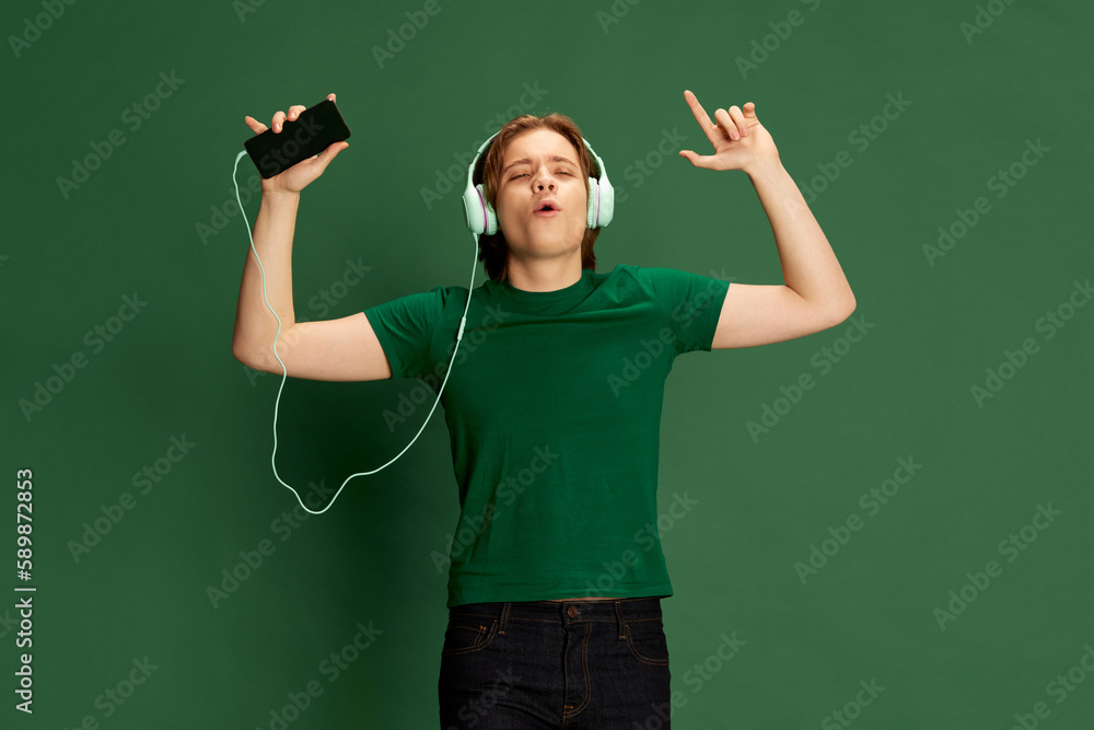 One handsome man, guy wearing headphones listening music with pleasure and singing over green background. Concept of music, hobby, human emotions, mood, ad