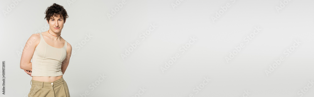 brunette pansexual model in tank top posing with hands behind back isolated on grey, banner. brunette pansexual model in tank top posing with hands behind back isolated on grey, banner.