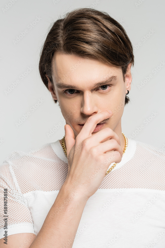 young pansexual person in white t-shirt looking at camera while holding hand near face isolated on grey. young pansexual person in white t-shirt looking at camera while holding hand near face isolated on grey.