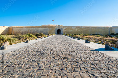 Grand entrance of the majestic fortress Fortaleza de Sagres showcased in a wide-angle shot, featuring a vast driveway leading to the giant gate and impressive wall under blue sky in Sagres, Portugal.