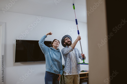 A young couple is looking at the ceiling and screaming while holding a mop because the upstairs neighbor is having a party with loud music or doing repairs in the apartment, workers are drilling with