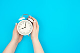 White alarm clock in female hands on a blue background, top view, concept of time management, deadline and work in the office.