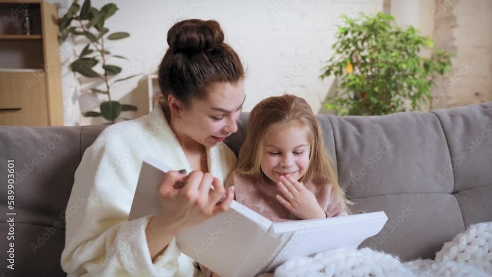 Young woman sitting on sofa with her little cute girl, daughter and reading books, fairy tales at home on daytime. Smiles, happiness. Family relationship, motherhood, childhood, love, caring concept Young woman sitting on sofa with her little cute girl, daughter and reading books, fairy tales at home on daytime. Smiles, happiness. Family relationship, motherhood, childhood, love, caring concept