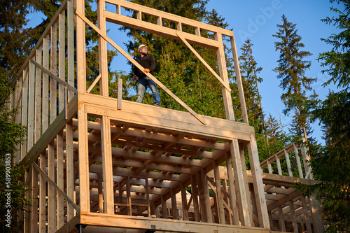 Carpenter constructing wooden framed two-story house near the forest. Bearded man with glasses holds hammer, dressed in protective helmet. Concept of environmentally friendly modern construction.