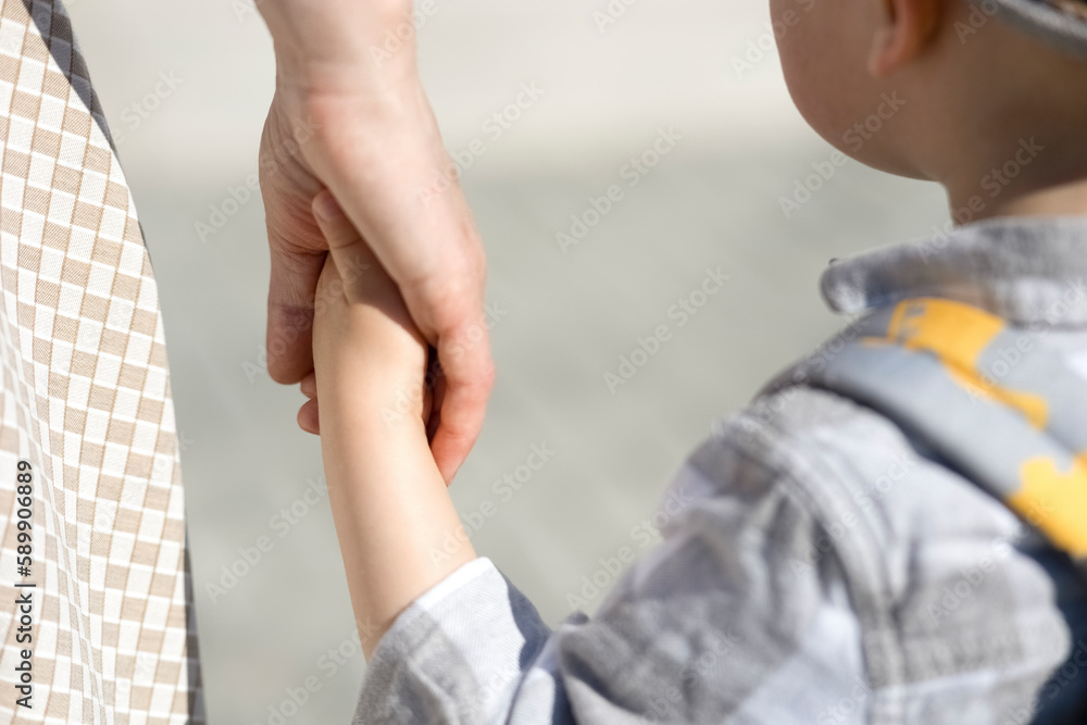 A woman holds a small child by the hand. A little boy in a gray hat and with a backpack on his back, holding his mother's hand. Mom takes the child to school, for a walk, to a children's camp or