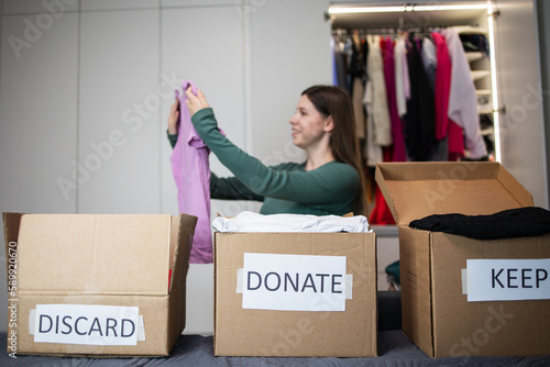 Unrecognizable woman sorts through her wardrobe. Three craft cardboard boxes of keep, discard, donation, donate. Charity clothing donations, help low income families, recycling, declutter, sustainable photo