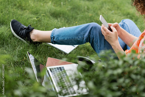 cropped view of young woman using smartphone while sitting near laptop and notebook on grass.