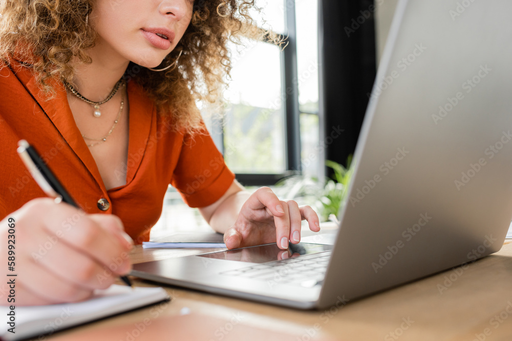 partial view of curly businesswoman taking notes in notebook while using laptop. partial view of curly businesswoman taking notes in notebook while using laptop.